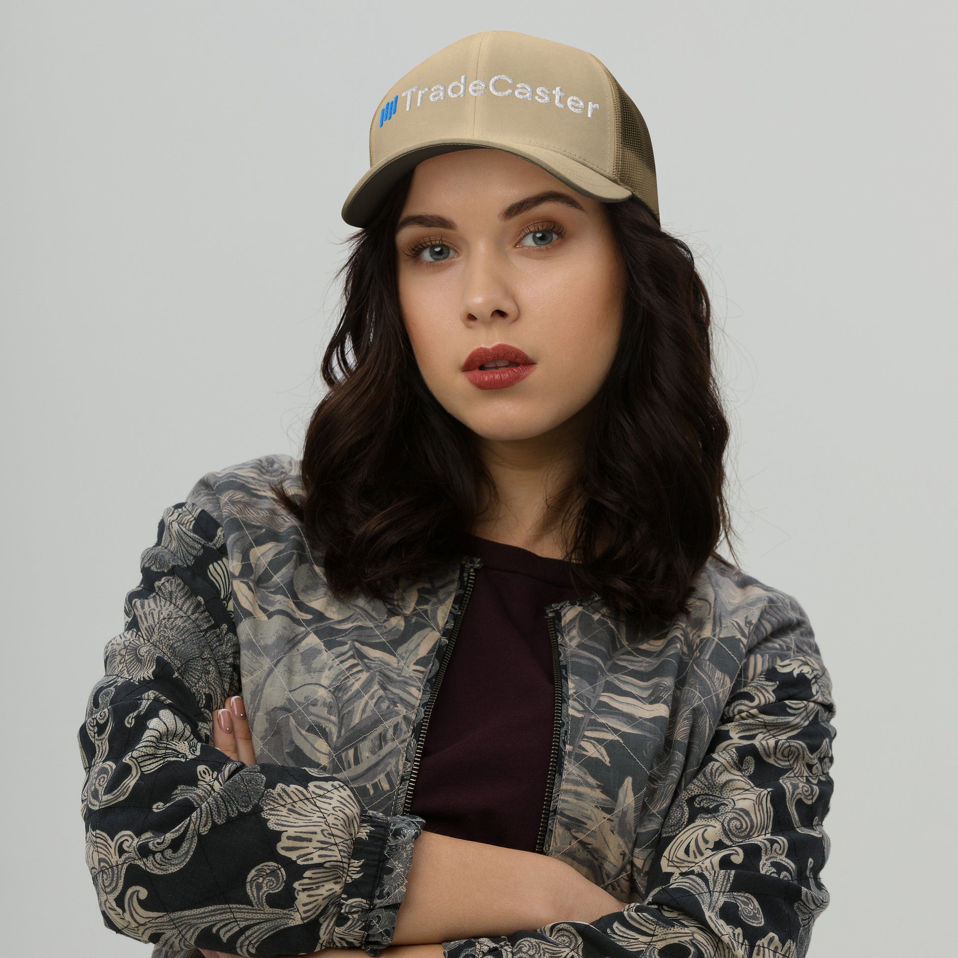 Woman rocking caramel TradeCaster trucker cap front view, arms crossed in gray jacket, white "TradeCaster" embroidery with blue logo