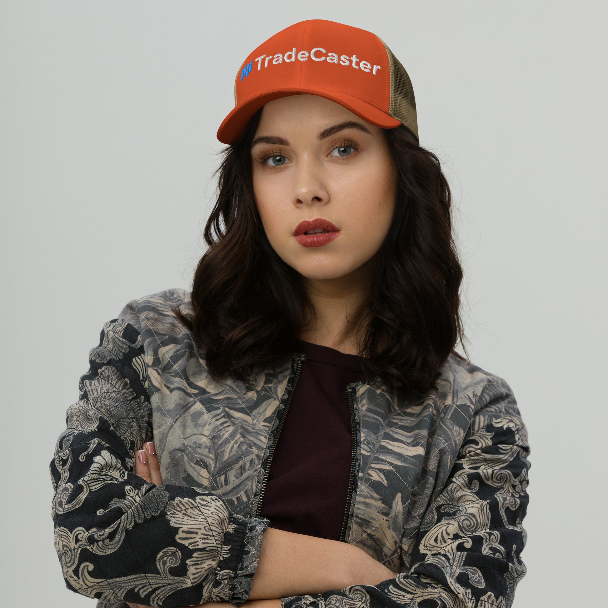 Fiery orange TradeCaster cap front on woman, arms crossed in gray jacket, camo mesh back peeks