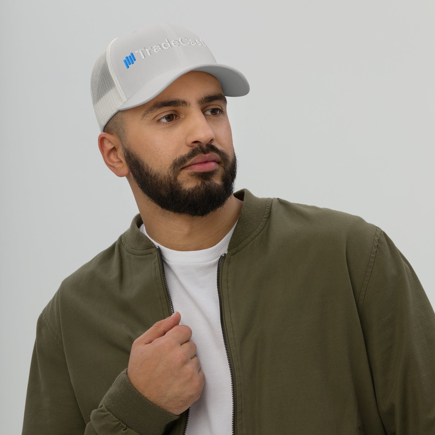 Side view bearded dude in dark heather gray TradeCaster cap, hand on face white tee olive jacket