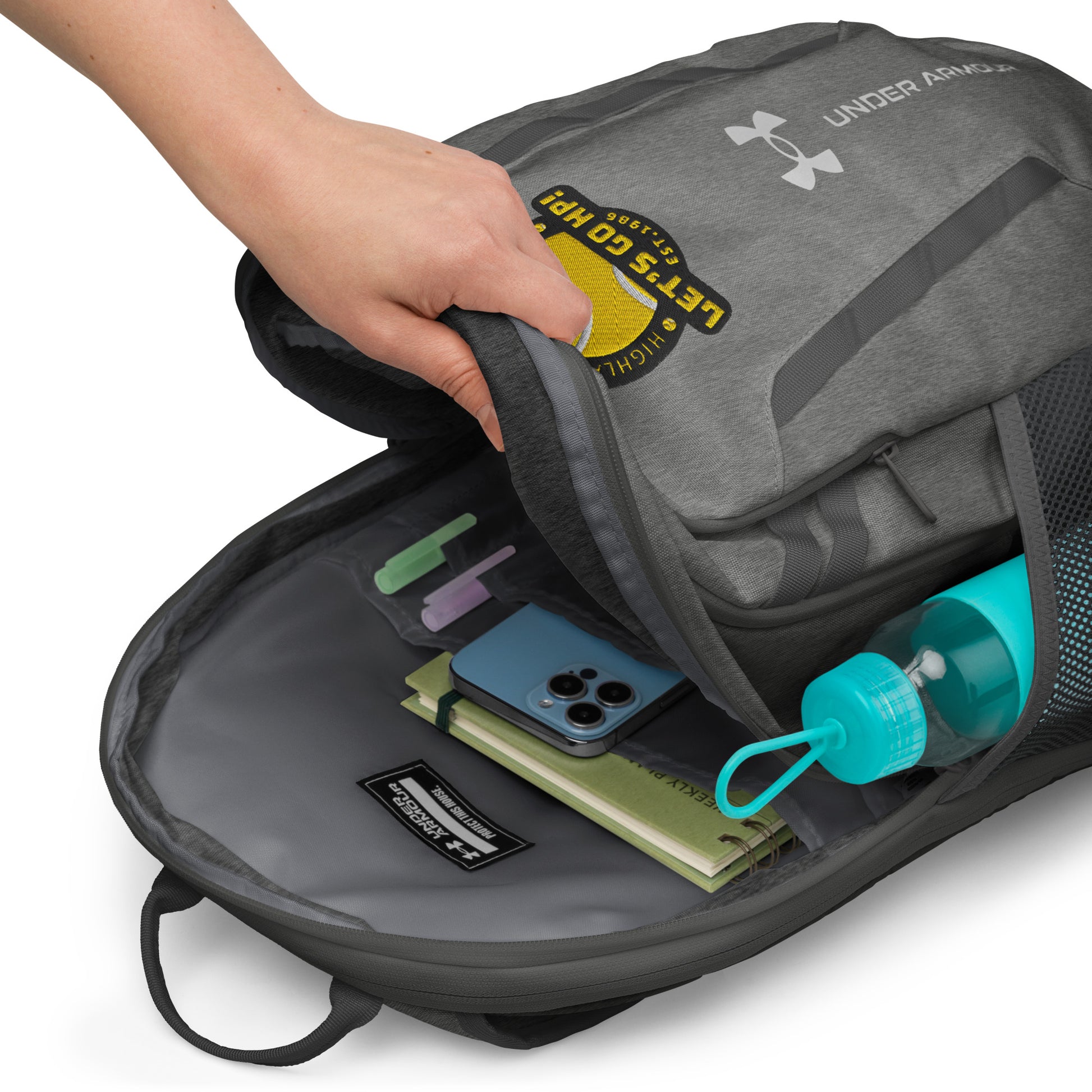 Hand opening heather gray Under Armour backpack, revealing gray interior with notebook, phone, and green water bottle in mesh pocket
