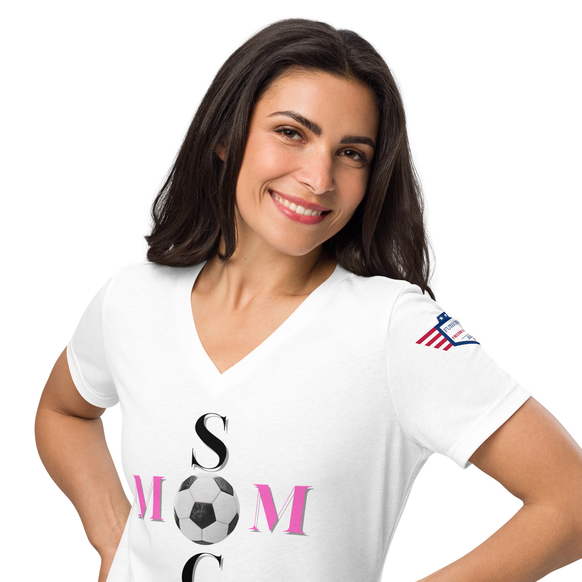 Smiling brunette model in white v-neck tee with "SOCCER MOM" black vertical print, pink "MOM" soccer ball graphic, flag sleeve patch, hands on hips